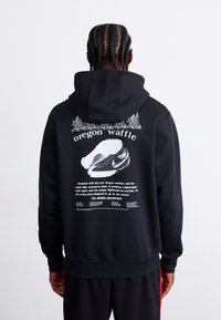 Sort hættetrøje med et stort grafisk design på ryggen, der inkluderer tekst og en illustration af en sko. Blødt materiale med en afslappet pasform.