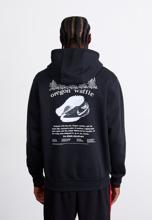 Sort hættetrøje med et stort grafisk design på ryggen, der inkluderer tekst og en illustration af en sko. Blødt materiale med en afslappet pasform.