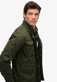 Veste vert olive avec un col haut, plusieurs poches avant et un tissu texturé. Portée sur une chemise à carreaux, avec un détail en patch.