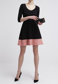 Robe noire et rose avec un corsage ajusté, des manches trois-quarts et une jupe évasée. Pochette noire texturée et escarpins noirs.
