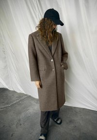 Bruine wollen jas met een getailleerd ontwerp, voorzien van een reverskraag, sluiting met twee knopen en zijzakken, gecombineerd met een zwarte pet en broek.