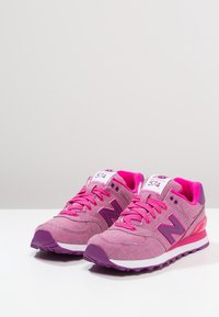 Ett par rosa New Balance 574 sneakers med lila detaljer och vita mellansålar på vit yta mot ljusgrå bakgrund.