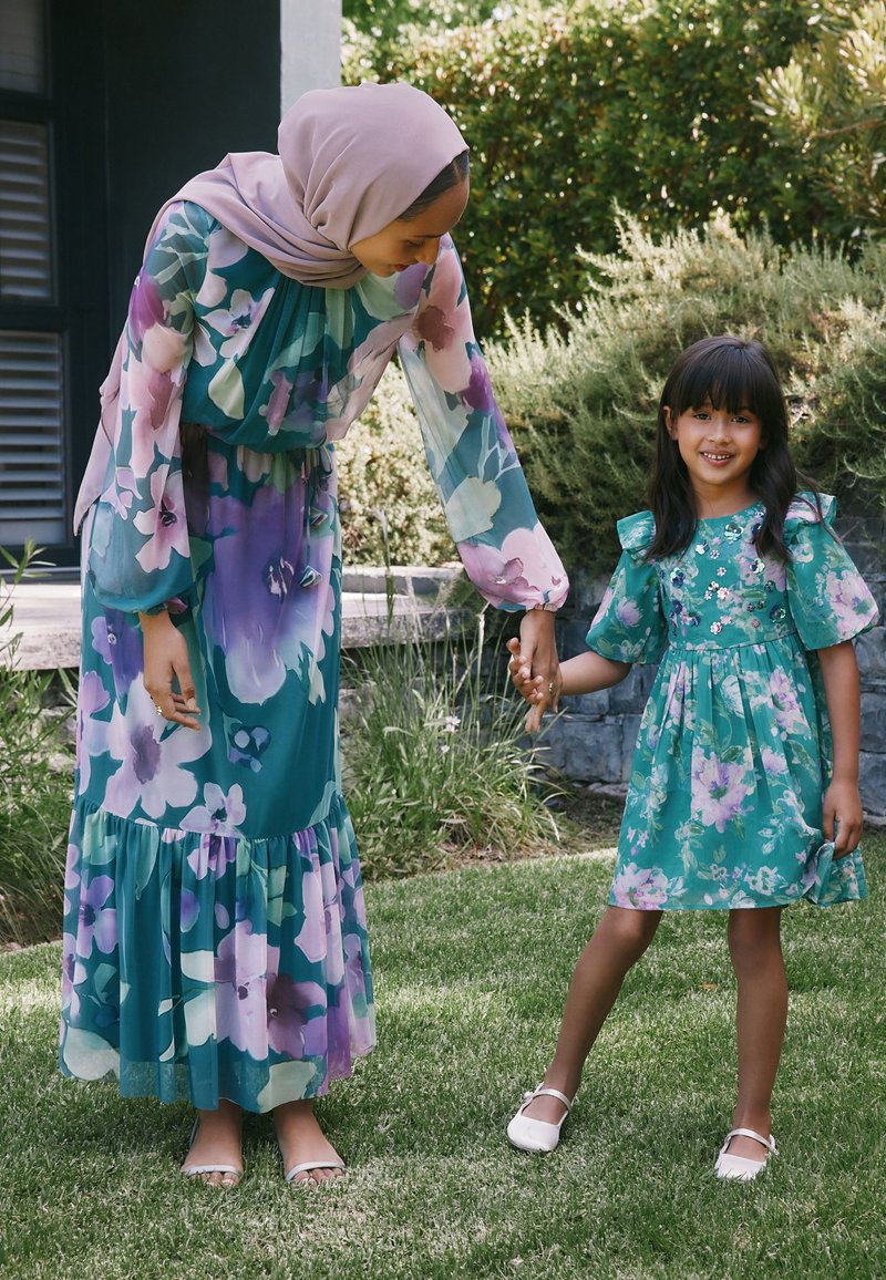 Mujer con pañuelo en la cabeza y vestido floral se inclina para tomar de la mano a una niña sonriente con vestido floral, que está de pie sobre el césped al aire libre.