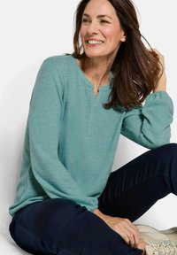 Un top lungo teal con scollo a V e polsini arricciati, abbinato a pantaloni scuri e scarpe chiare. Tessuto morbido, design casual.