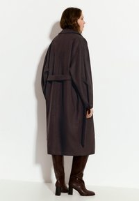 Lang brun uldfrakke med bælte i taljen, kravehals og dropped skuldre, parret med knæhøje brune læderstøvler.