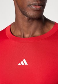 Rød træningsshirt med rund hals, lavet af glat materiale, med et hvidt Adidas-logo på brystet. Syningerne er rene og enkle.