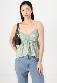 Grønn bluse med blomsterdesign, tynne stropper, samlet kant og blondebesetning. Kombinert med lyseblå høytlivsjeans og en mørk håndveske.