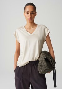 Beige v-hals t-shirt met korte mouwen, gemaakt van zachte stof. Gecombineerd met donkere, loszittende broek en een geweven groene tas.