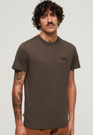 Camiseta de algodón verde oscuro con cuello redondo, mangas cortas y logo bordado. Tatuajes notables visibles en el brazo.