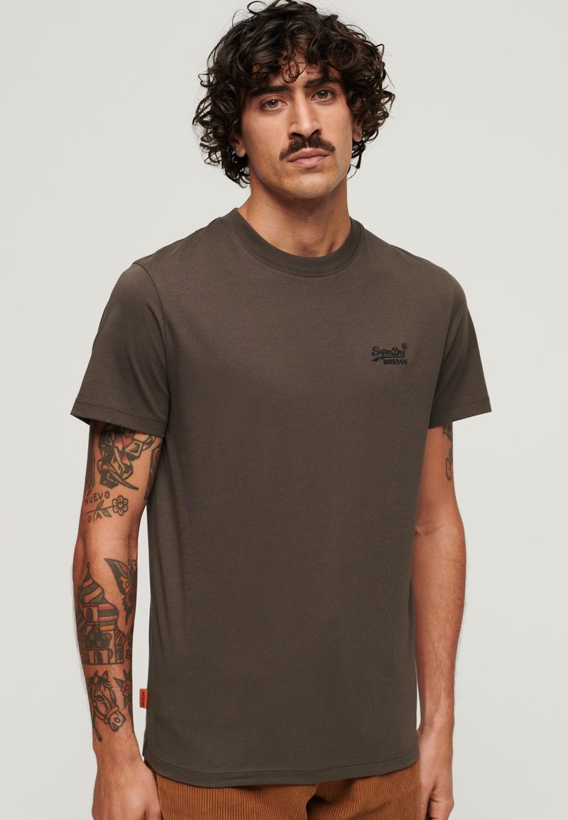 Camiseta de algodón verde oscuro con cuello redondo, mangas cortas y logo bordado. Tatuajes notables visibles en el brazo.