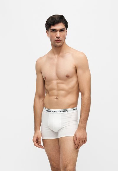 Мъж без риза с тъмна коса, носещ бели боксерки Polo Ralph Lauren, стои на бял фон.