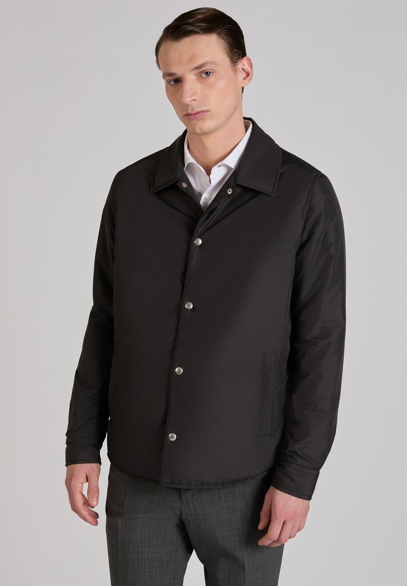 Mężczyzna w czarnej kurtce zapinanej na guziki, białej koszuli i szarych spodniach w kratę, stojący na tle jednolitego, jasnoszarego tła.