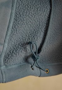 Primo piano di tessuto in pile blu con texture morbida visibile e coulisse elastica regolabile con blocco di plastica e occhielli metallici.