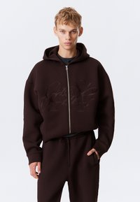 Mørkebrun zip-up hettegenser i myk stoff med en avslappet passform, med et preget design på brystet og kengurulommer.