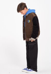Brązowa kurtka varsity z kordurowego materiału i czarnej tkaniny z niebieską bluzą z kapturem, z detalem logo i paskowanymi mankietami, zestawiona z czarnymi spodniami i niebieskimi trampkami.