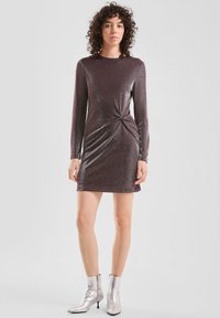 Kobieta z kręconymi włosami nosi błyszczącą fioletową mini sukienkę z długim rękawem, gromadzoną z boku, oraz srebrne botki za kostkę, stojąc na jednolitym tle.