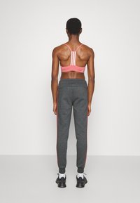 Persona vestida con un sujetador deportivo rosa, pantalones jogger gris oscuro con franjas rojas, calcetines blancos y zapatillas negras, de pie frente a una pared blanca.