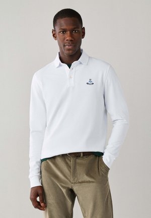 Hombre con camisa polo blanca de manga larga con pequeño logo en el pecho y pantalones de pana beige, de pie con una mano en el bolsillo.