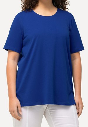 Personne portant une chemise bleue unie à manches courtes et un pantalon blanc, visible du cou jusqu'au milieu des cuisses sur un fond uni.