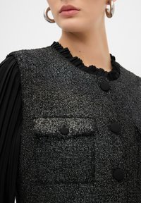Chaleco negro texturizado con un sutil brillo, con un cuello volado, dos bolsillos frontales y cierres de botón negros.