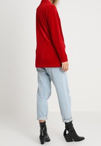 Haut rouge à manches longues en velours avec col, associé à un jean denim bleu clair retroussé. Bottines noires avec un détail brillant.