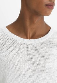 Bílý pletený svetr s volným, otevřeným vzorem. Má kulatý výstřih a žebrované manžety, které zdůrazňují měkkou, lehkou texturu.