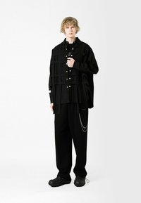Camisa negra de corte oversize con estampado de cuadros y botones plateados, combinada con pantalones holgados negros con un acento de cadena y calzado negro texturizado.