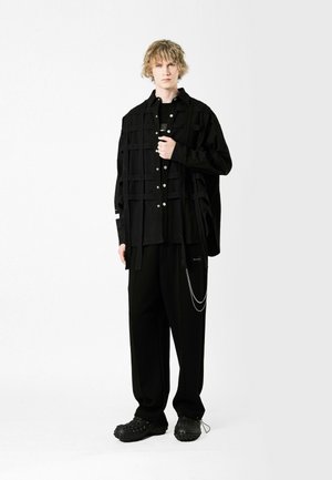 Camisa negra de corte oversize con estampado de cuadros y botones plateados, combinada con pantalones holgados negros con un acento de cadena y calzado negro texturizado.
