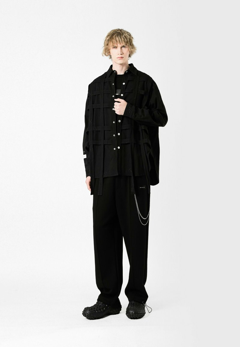 Camisa negra de corte oversize con estampado de cuadros y botones plateados, combinada con pantalones holgados negros con un acento de cadena y calzado negro texturizado.
