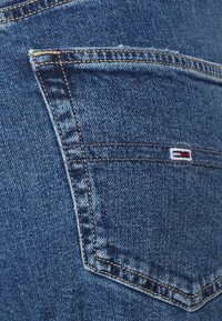 Bolsillo trasero de unos vaqueros de mezclilla, en azul con costura visible. Presenta un pequeño logotipo bordado con franjas rojas, blancas y azules. Tela texturada.