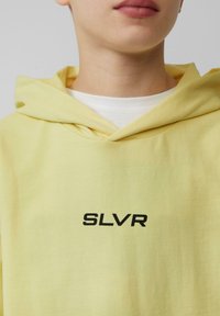 Żółta hoodie z kapturem, wykonana z miękkiego materiału, z czarnym logo "SLVR" z przodu, z warstwą białej koszuli pod spodem.