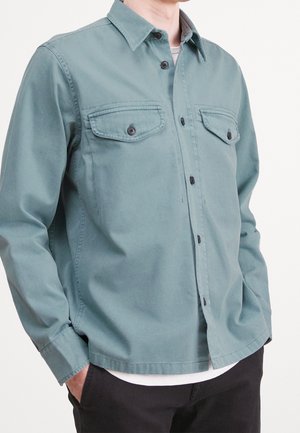Man draagt een turquoise blouse met knopen en twee met knopen gesloten borstzakken over een wit hemd, handen in de zakken van zwarte broek.