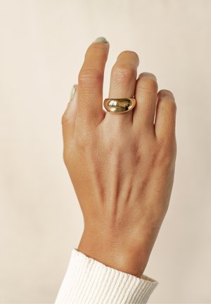 Goldfarbener Ring mit einer glatten, reflektierenden Oberfläche, präsentiert auf einer Hand mit heller Haut. Die Hand trägt einen strukturierten weißen Pullover.