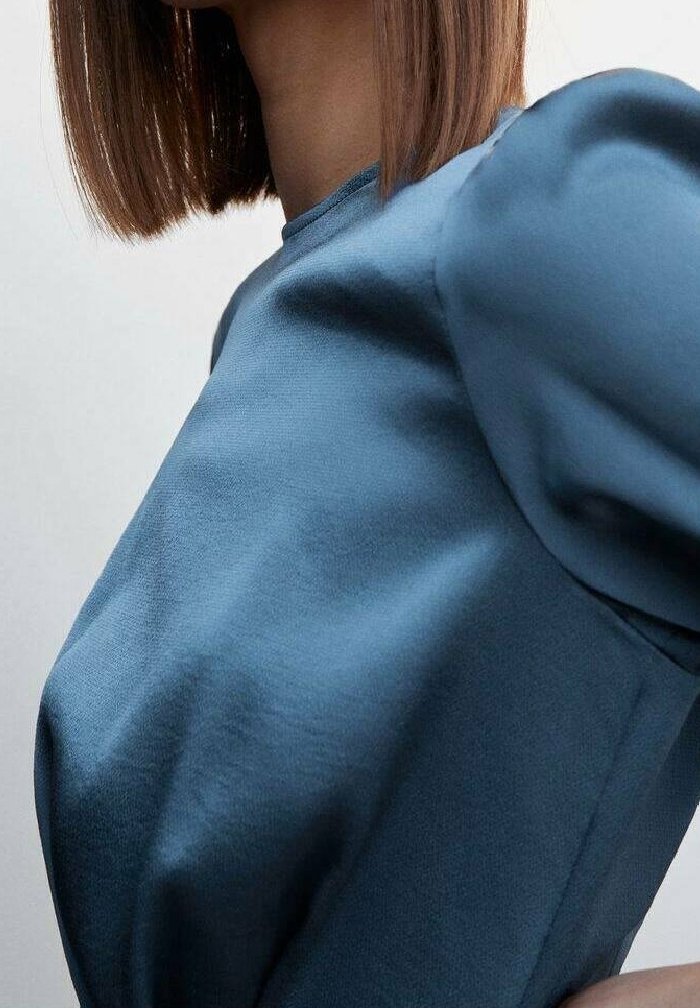 Femme portant un haut bleu à manches bouffantes, visible des épaules jusqu'au milieu du torse avec des cheveux bruns raides visibles.