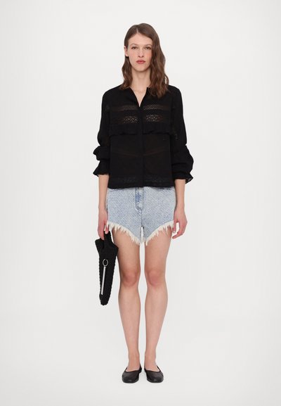 Schwarze durchsichtige Bluse mit Spitzenverzierungen, Rüschensleeves, kombiniert mit hellblauen, ausgefransten Denim-Shorts und schwarzen Ballerinas, hält eine schwarze Clutch.