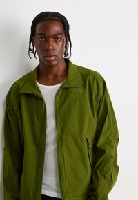 Young man with braided hair wearing a green North Face jacket over a white ribbed shirt against a plain light background.