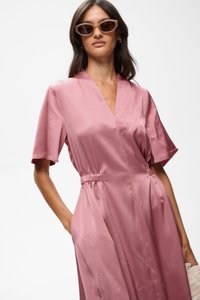 Femme portant des lunettes de soleil œil de chat et une robe portefeuille rose poudré texturée avec manches courtes et taille élastique, tenant une pochette tissée.