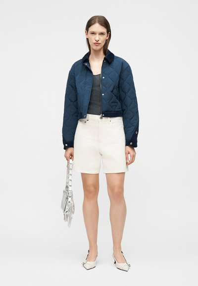 Femme portant une veste matelassée bleu marine sur un haut gris foncé, un short blanc et des talons pointus blancs, tenant un petit sac à main argenté.