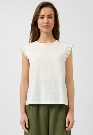 Femme portant un haut blanc sans manches avec une bordure en dentelle aux manches et un pantalon vert olive, debout devant un fond uni.