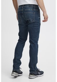 Vaqueros de mezclilla azul oscuro con corte recto, que cuentan con dos bolsillos traseros, costuras sutiles y una cintura de altura media. Llevados con zapatillas negras.