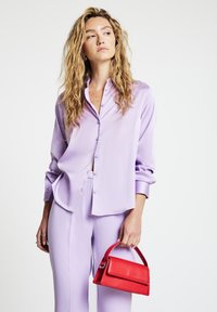 Woman with wavy blonde hair wearing a lavender button-up shirt and pants, holding a small red handbag by the handle.
