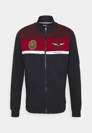 Herenjack met ritssluiting in zwart, bordeauxrood en rood kleurvlakken, logopatches op de borst en een zak met rits op de linker mouw.