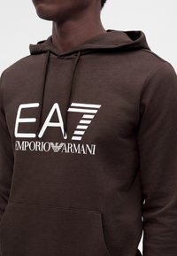 Άτομο που φοράει σκούρο καφέ φούτερ με κουκούλα με λευκό λογότυπο EA7 Emporio Armani τυπωμένο στο στήθος, με μπροστινή τσέπη και κουκούλα με κορδόνι.