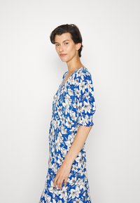 Vestido azul com padrões de lírios brancos, apresentando mangas curtas bufantes e um decote em V profundo. Tecido suave com um design fluido.