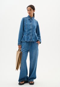 Blauwe denim peplum blouse met een kraag en gerimpelde zoom, gecombineerd met wijde denim broeken en een beige handtas. Platte sandalen.