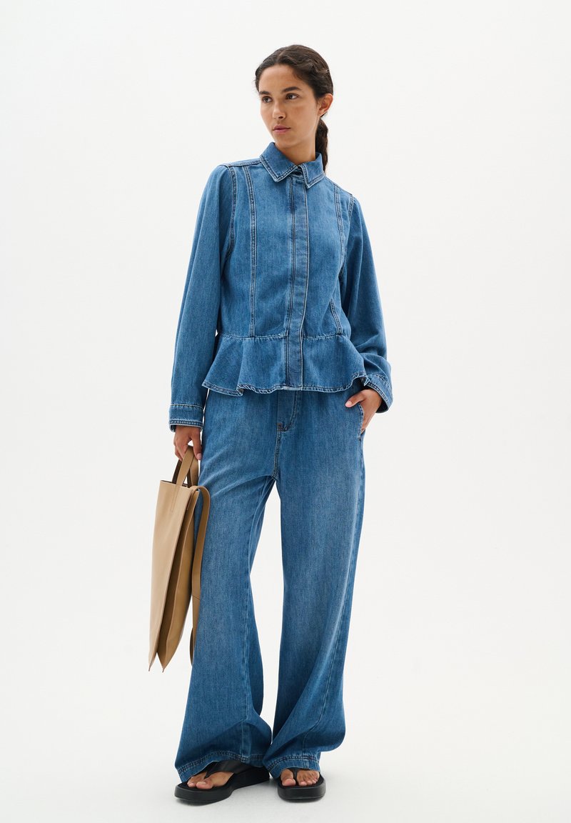 Blauwe denim peplum blouse met een kraag en gerimpelde zoom, gecombineerd met wijde denim broeken en een beige handtas. Platte sandalen.