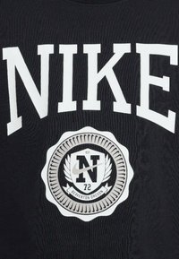 Camiseta negra con gran texto blanco "NIKE", un emblema circular con "N" y "72", y acentos de laurel en el centro. Tejido texturizado.