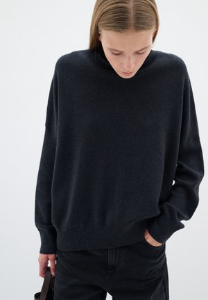 Dunkelgrauer, übergroßer Pullover mit hohem Kragen und überschnittenen Schultern. Weicher Stoff, gerippter Saum, kombiniert mit schwarzen Hosen und einer Handtasche.