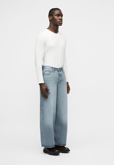 Νέος άνδρας που στέκεται, φοράει λευκό πουκάμισο με μακριά μανίκια, φαρδιά ανοιχτό μπλε τζιν και σκούρα καφέ παπούτσια σε απλό φόντο.