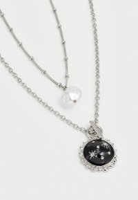 Collier en argent superposé présentant une perle blanche irrégulière et un pendentif noir rond avec des motifs d'étoiles et de petites pierres claires.